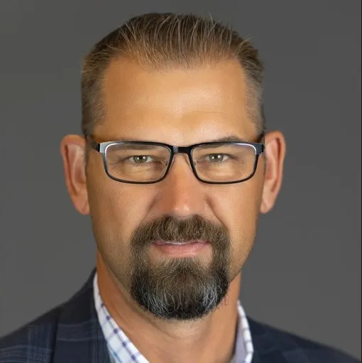 Middle-aged man with short hair, glasses, and a trimmed beard wearing a plaid jacket and checkered shirt against a gray background.