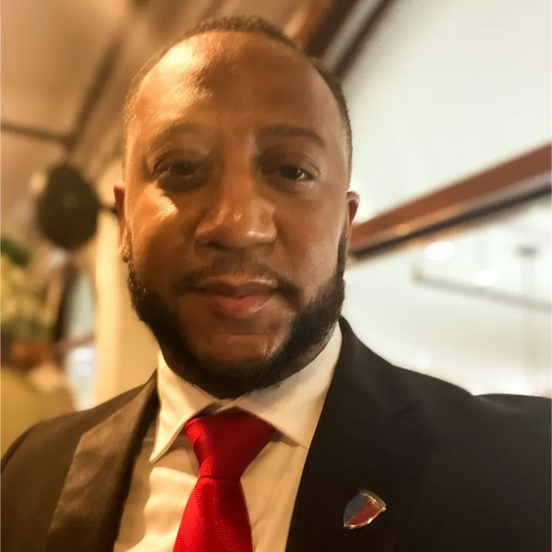 Headshot of a man with a beard wearing a black suit, white shirt, and red tie indoors.