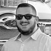 Man with short hair, beard, and sunglasses smiling in front of a large vehicle grill.