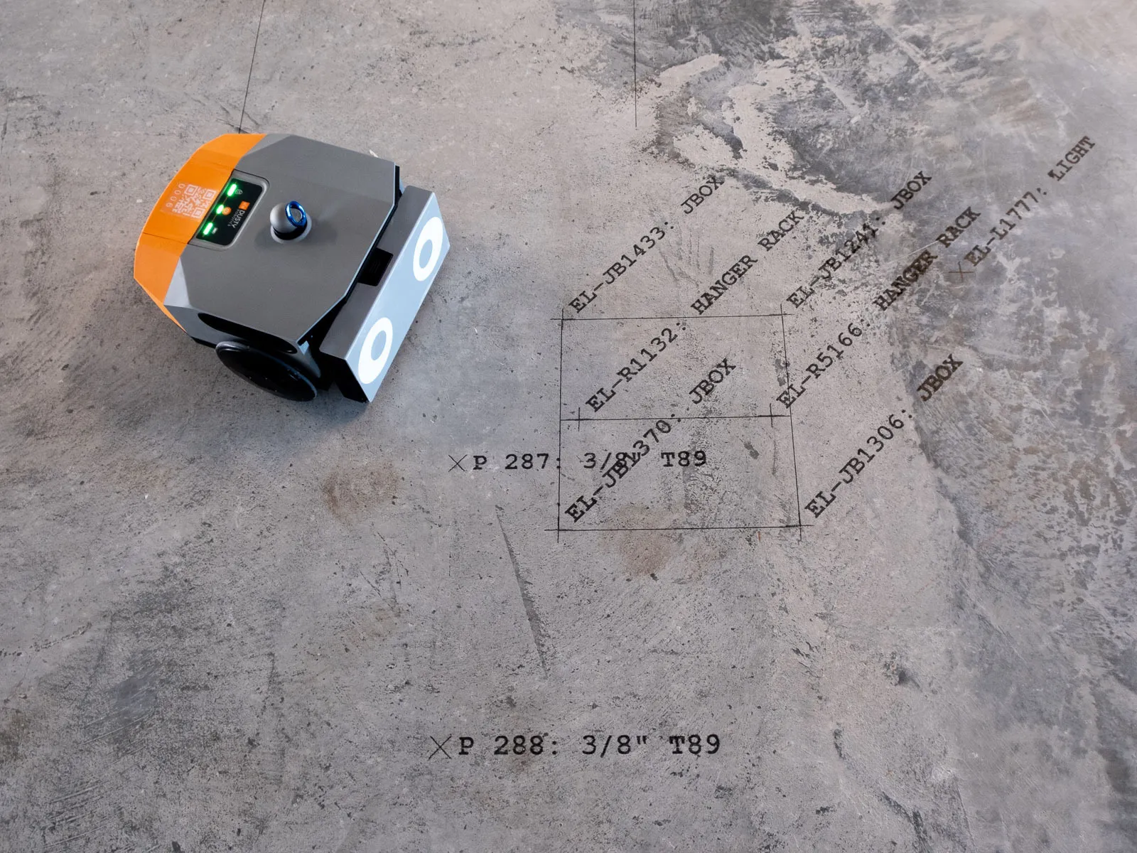 Small robotic device with lights and a knob on a concrete floor marked with black electrical layout symbols and text.