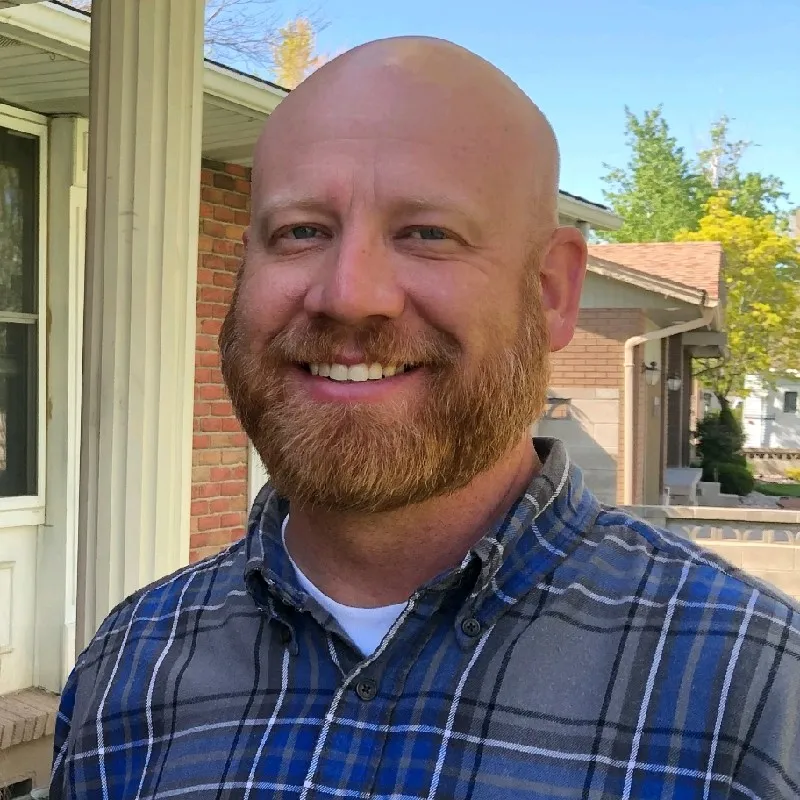 Travis Brock. Smiling man with a red beard wearing a blue plaid shirt outdoors in front of a brick house.