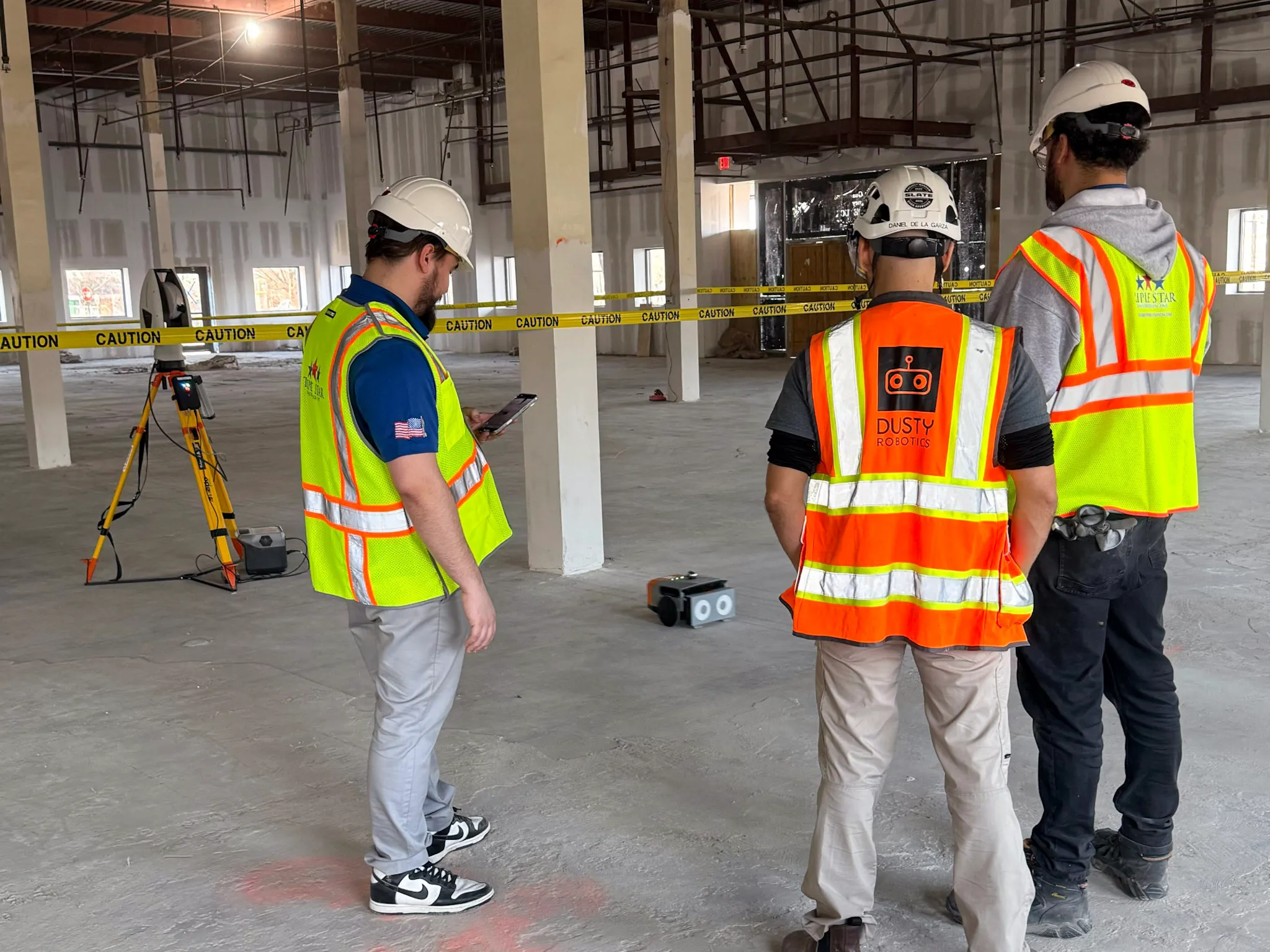 Three construction workers wearing hard hats and high-visibility vests standing inside a large unfinished building with caution tape and surveying equipment.