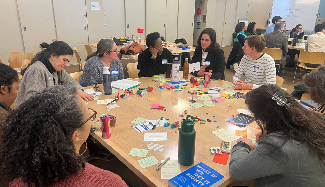 Group of adults collaborating in a workshop setting, gathered around tables with notes and materials while one person speaks and others listen.