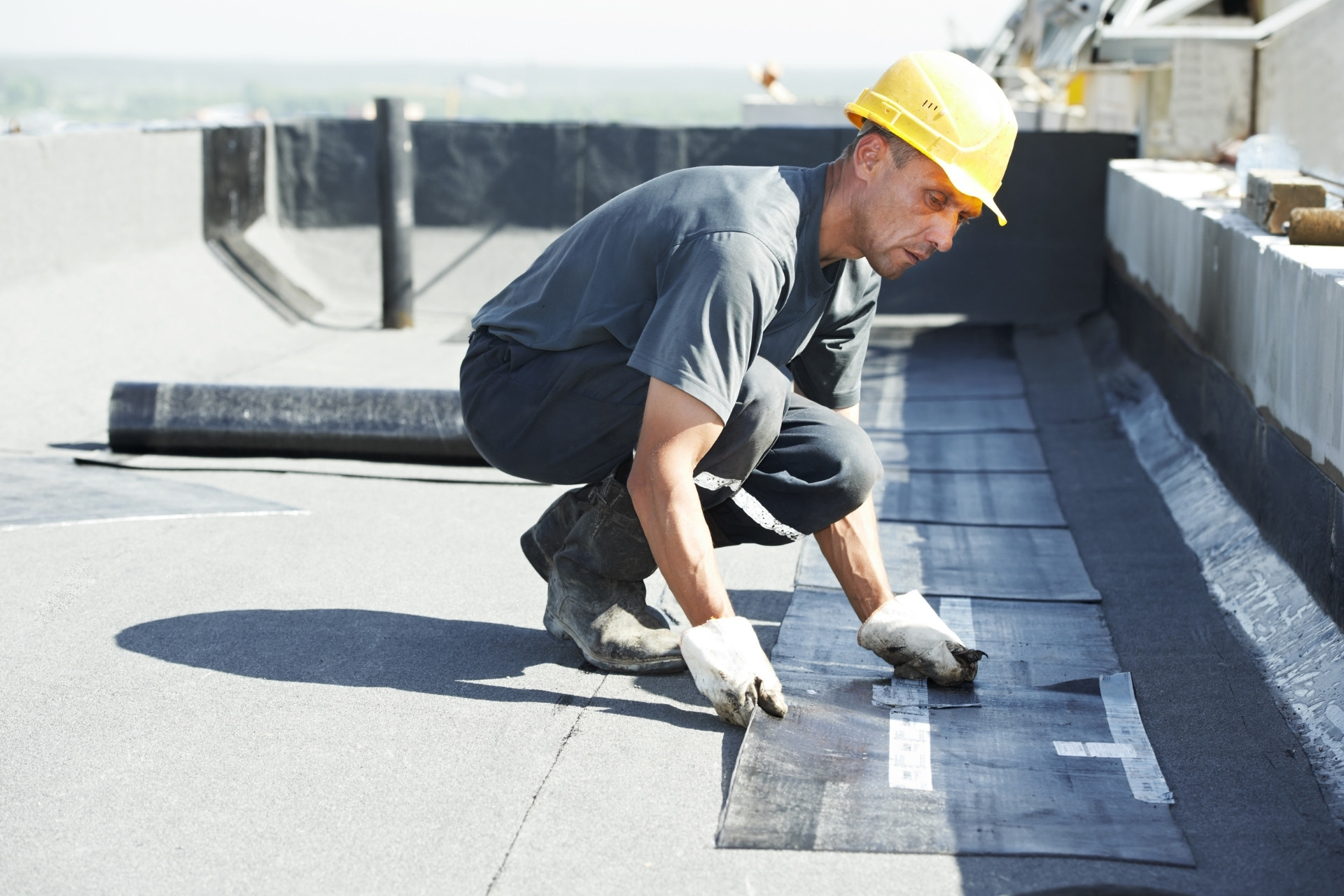 Construction worker in a yellow hard hat and gloves applying waterproof membrane sheets during a commercial roof repair.