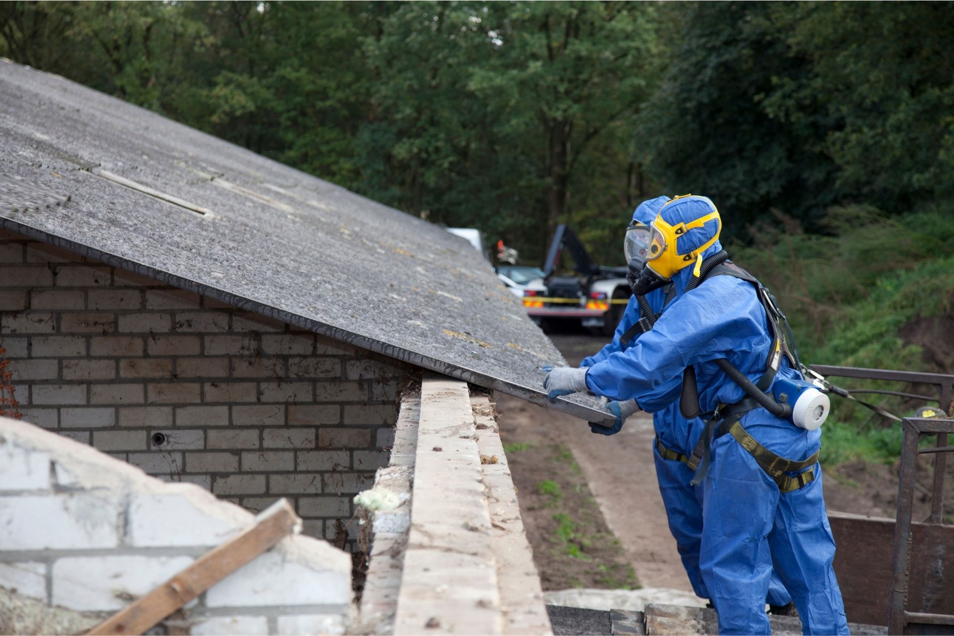 Two workers in full hazmat suits and respirators carefully remove an old asbestos roof sheet from a brick building, illustrating safety compliance with asbestos regulation during demolition.