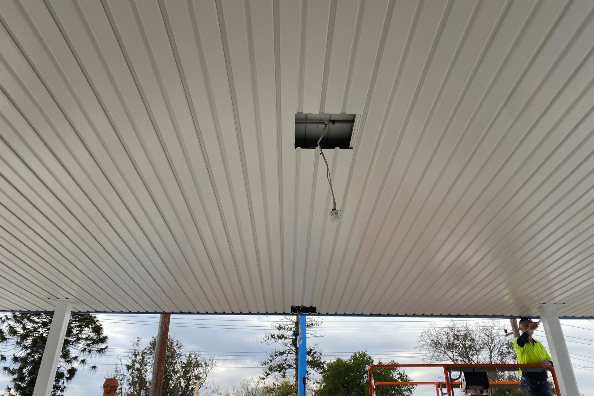 A close-up view of the underside of a newly installed white metal roofing canopy at a petrol station, with a cut-out section exposing wiring for lighting installation.