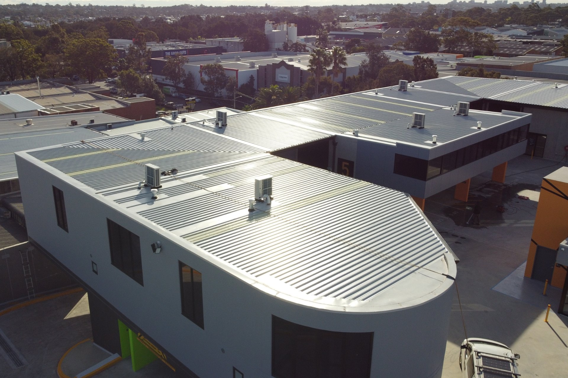 Aerial view of a commercial building complex featuring clean, new metal roofing with visible HVAC units and skylights, surrounded by an urban industrial area.