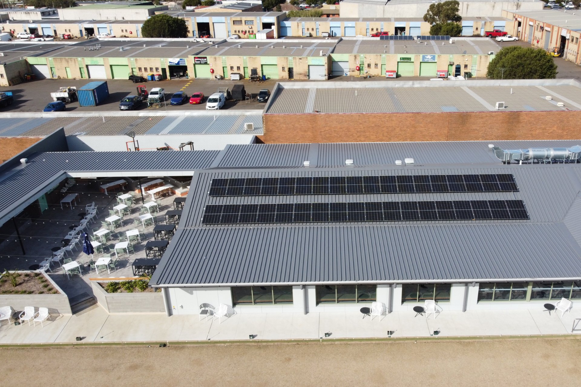 Commercial building with metal roofing and solar panels above an outdoor dining area with modern seating. Surrounded by older industrial units in the background.