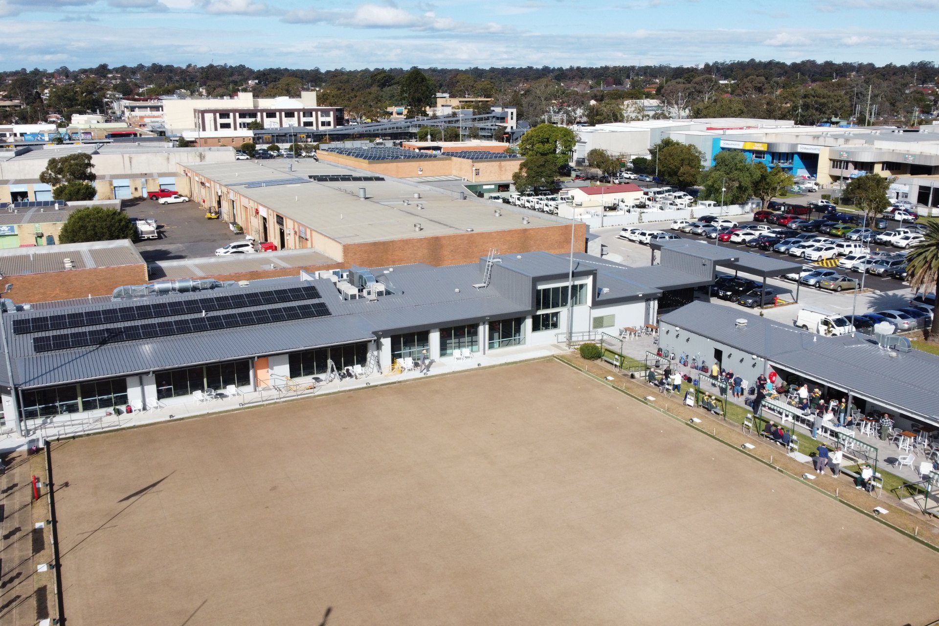 Commercial building with metal roofing and solar panels above an outdoor dining area with modern seating. Surrounded by older industrial units in the background.