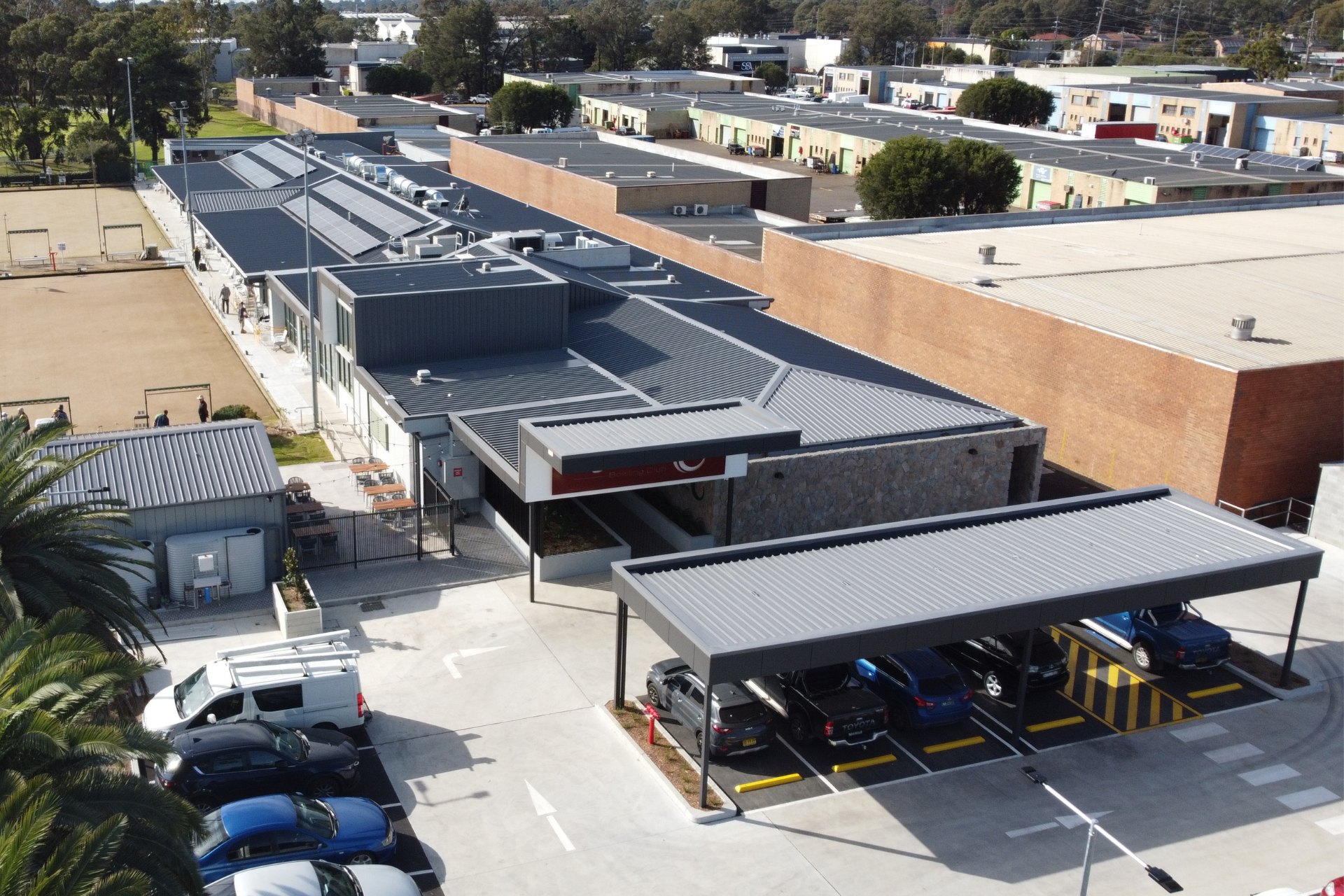 Commercial building with metal roofing and solar panels above an outdoor dining area with modern seating. Surrounded by older industrial units in the background.