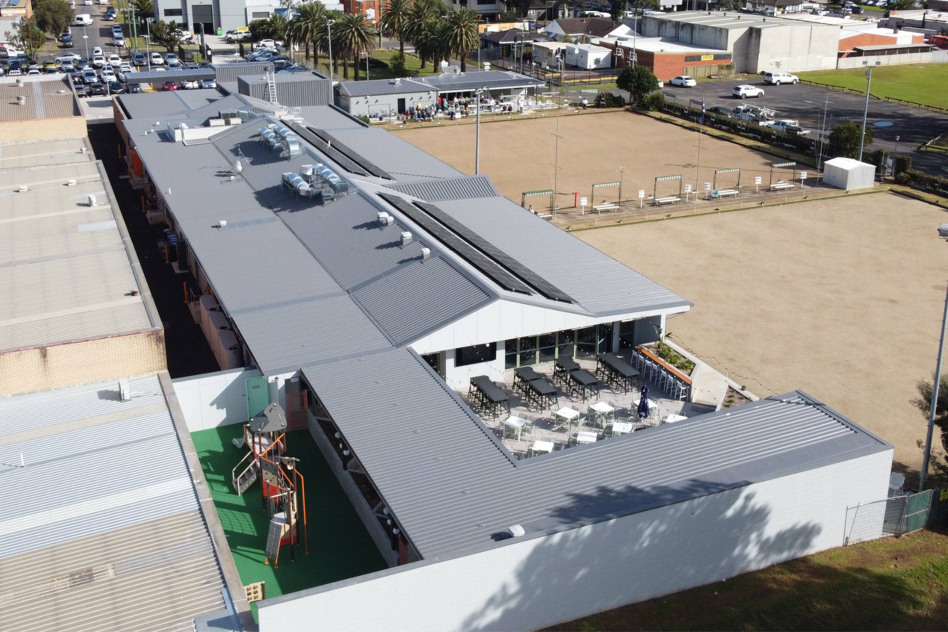 Commercial building with metal roofing and solar panels above an outdoor dining area with modern seating. Surrounded by older industrial units in the background.