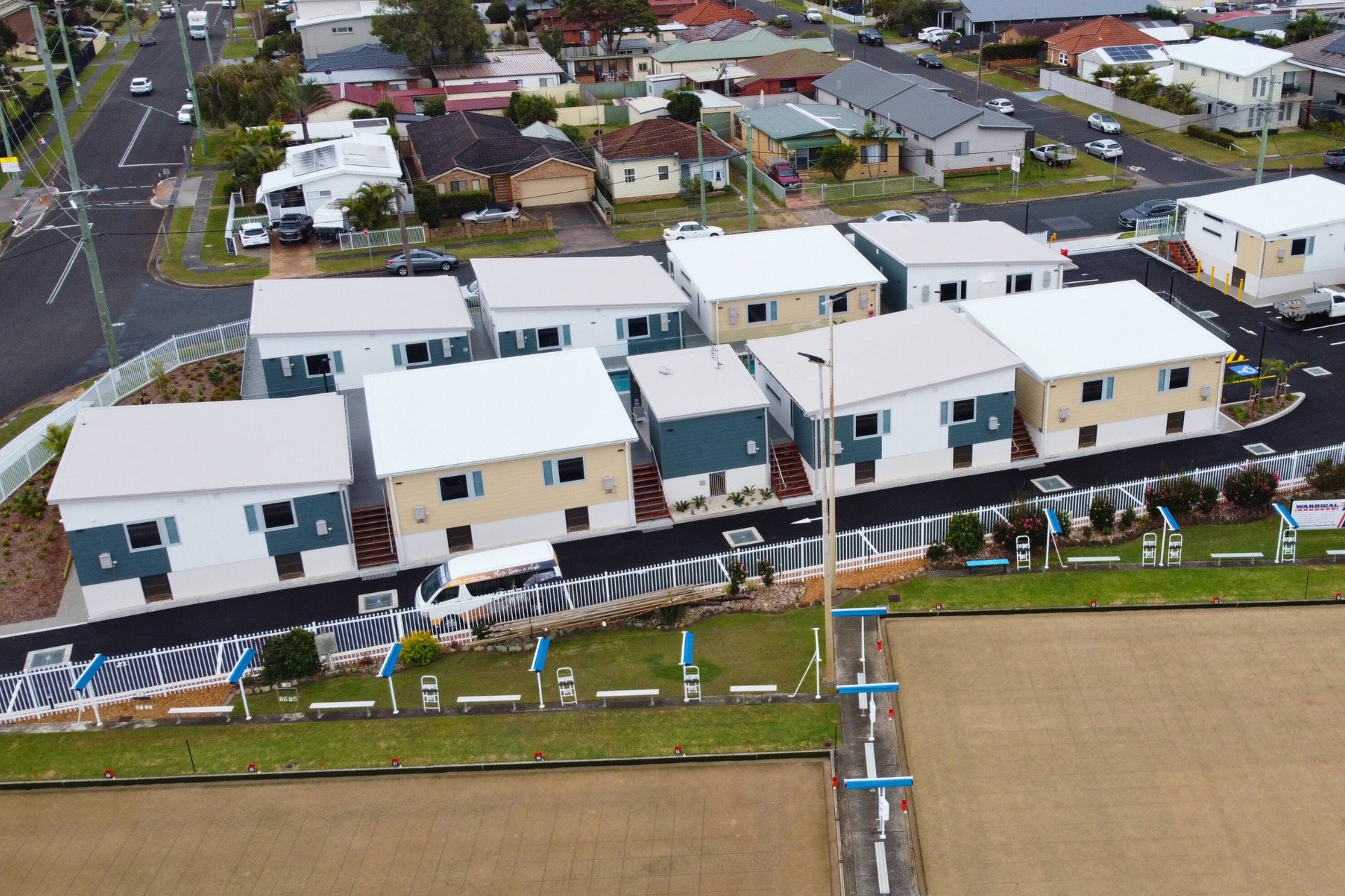 New motel development featuring rows of compact buildings with fresh white new commercial roofing. The clean, uniform roofs complement the modern exterior designs, set beside a lawn bowling green and residential neighborhood.
