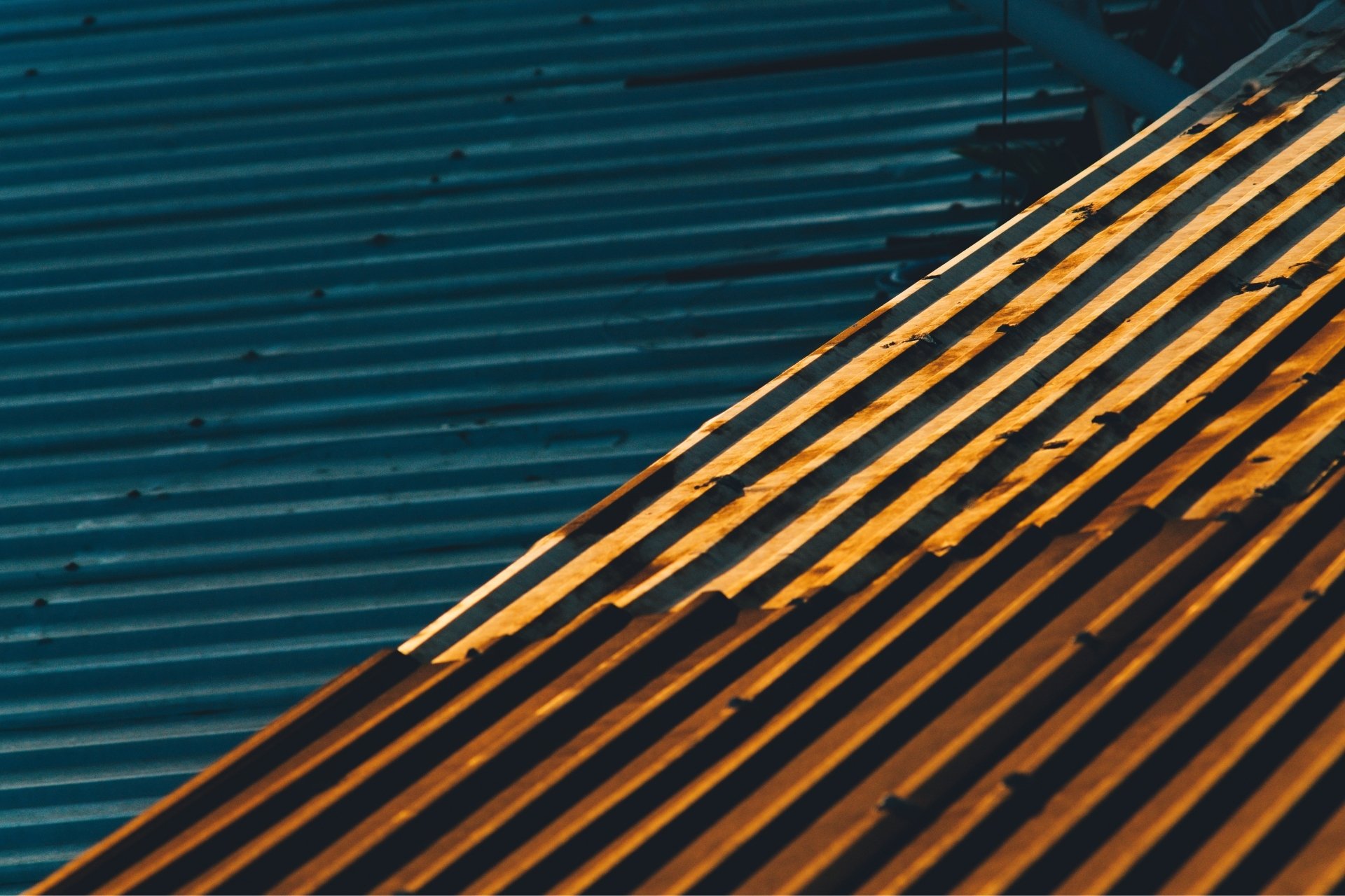 Close-up view of metal roofing panels with alternating shadows and sunlight casting bold diagonal lines across the surface. The image emphasizes the texture, durability, and clean lines associated with modern roofing solutions.