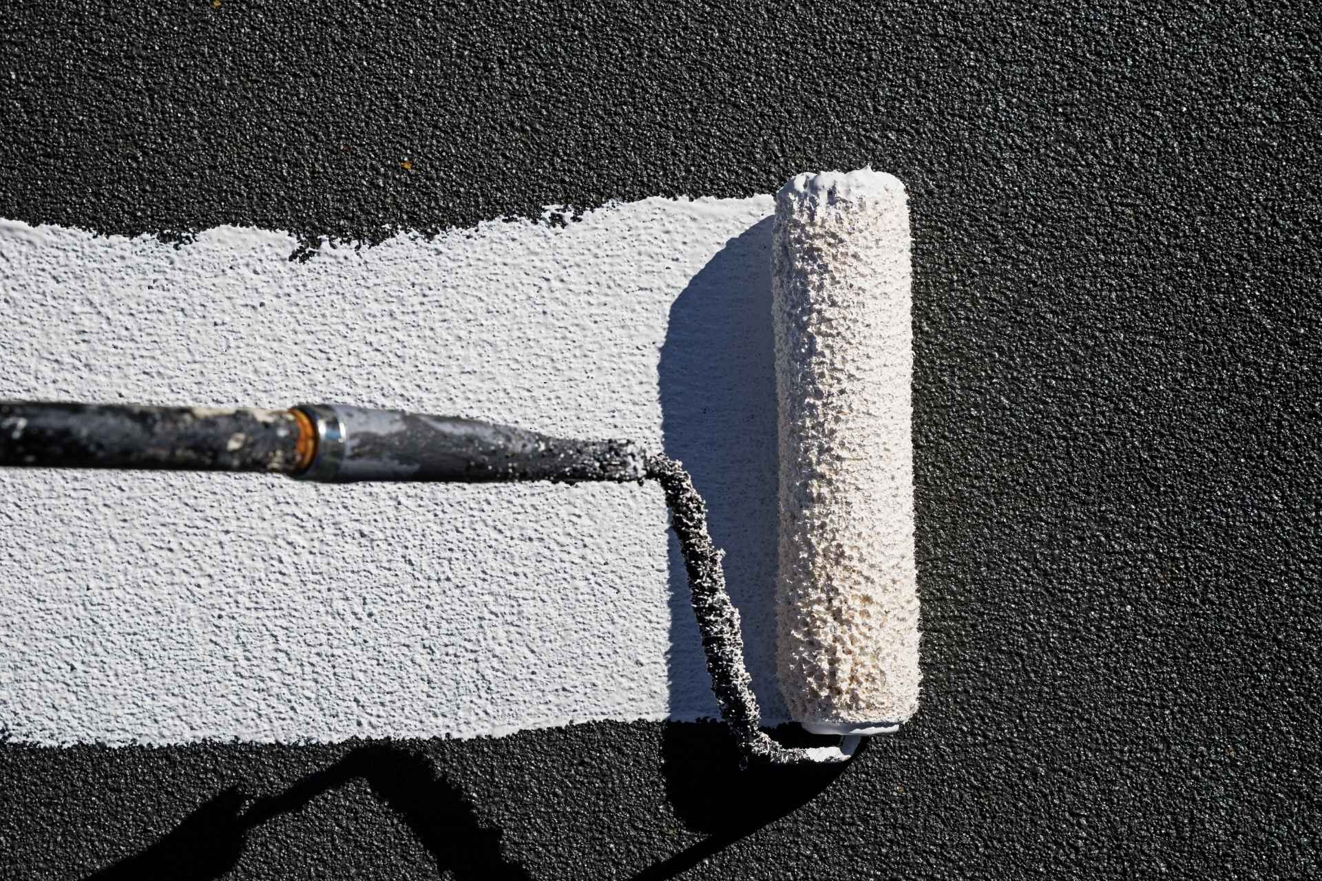 Close-up of a textured roller applying a thick white roof coating over a black surface. The image shows the contrast between coated and uncoated areas, highlighting the protective layer being applied during a roofing maintenance process.