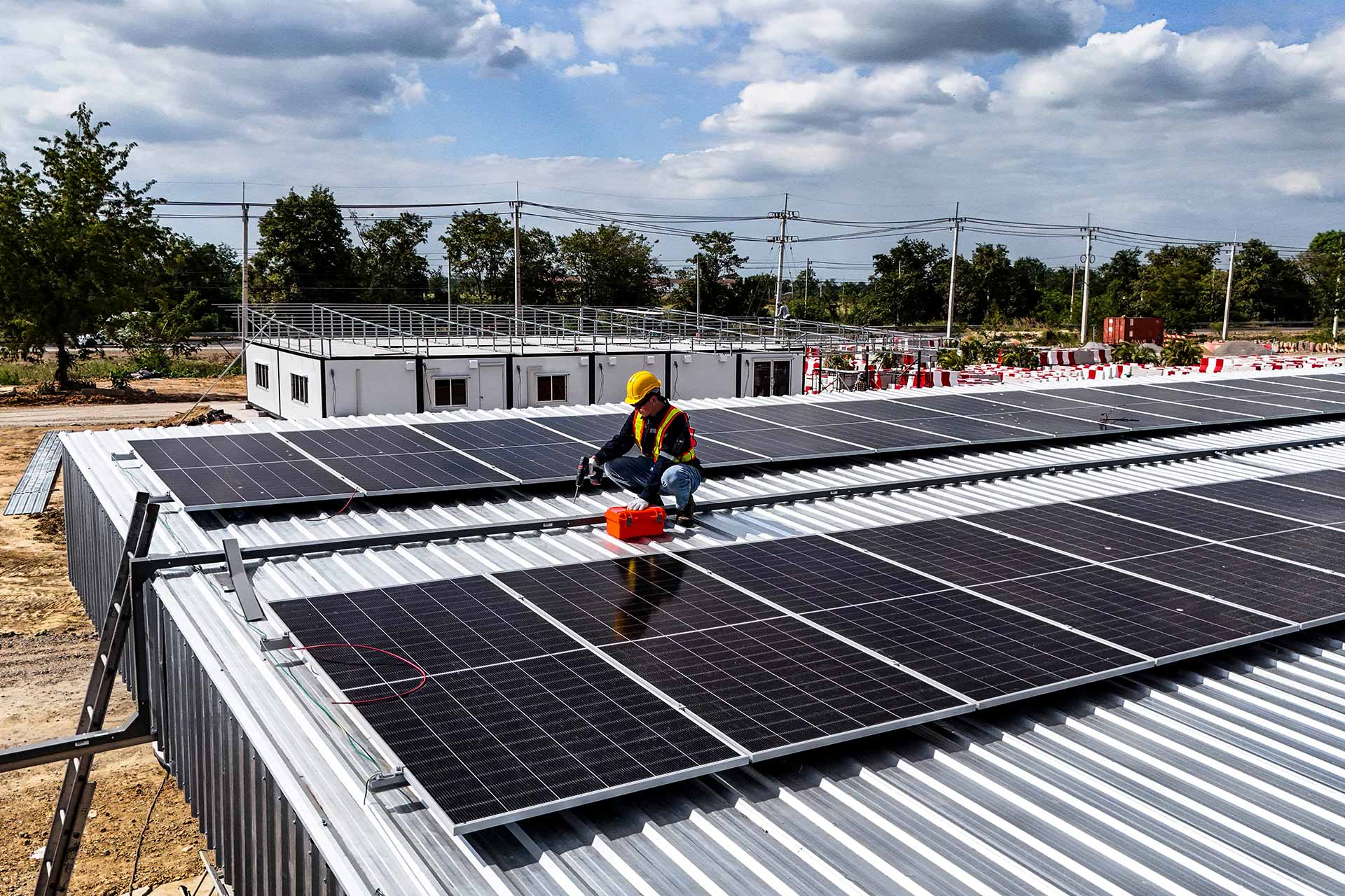 A worker in a yellow hard hat and safety vest installs solar PV panels on a corrugated metal roof.