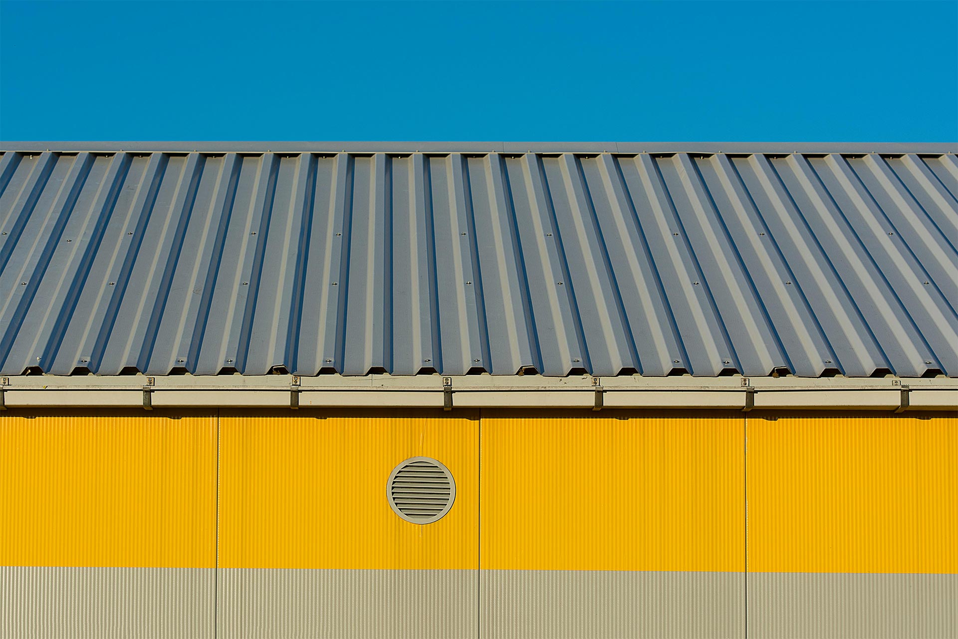 Close-up of a gray metal roof system on a yellow industrial building with a round wall vent and clear blue sky.