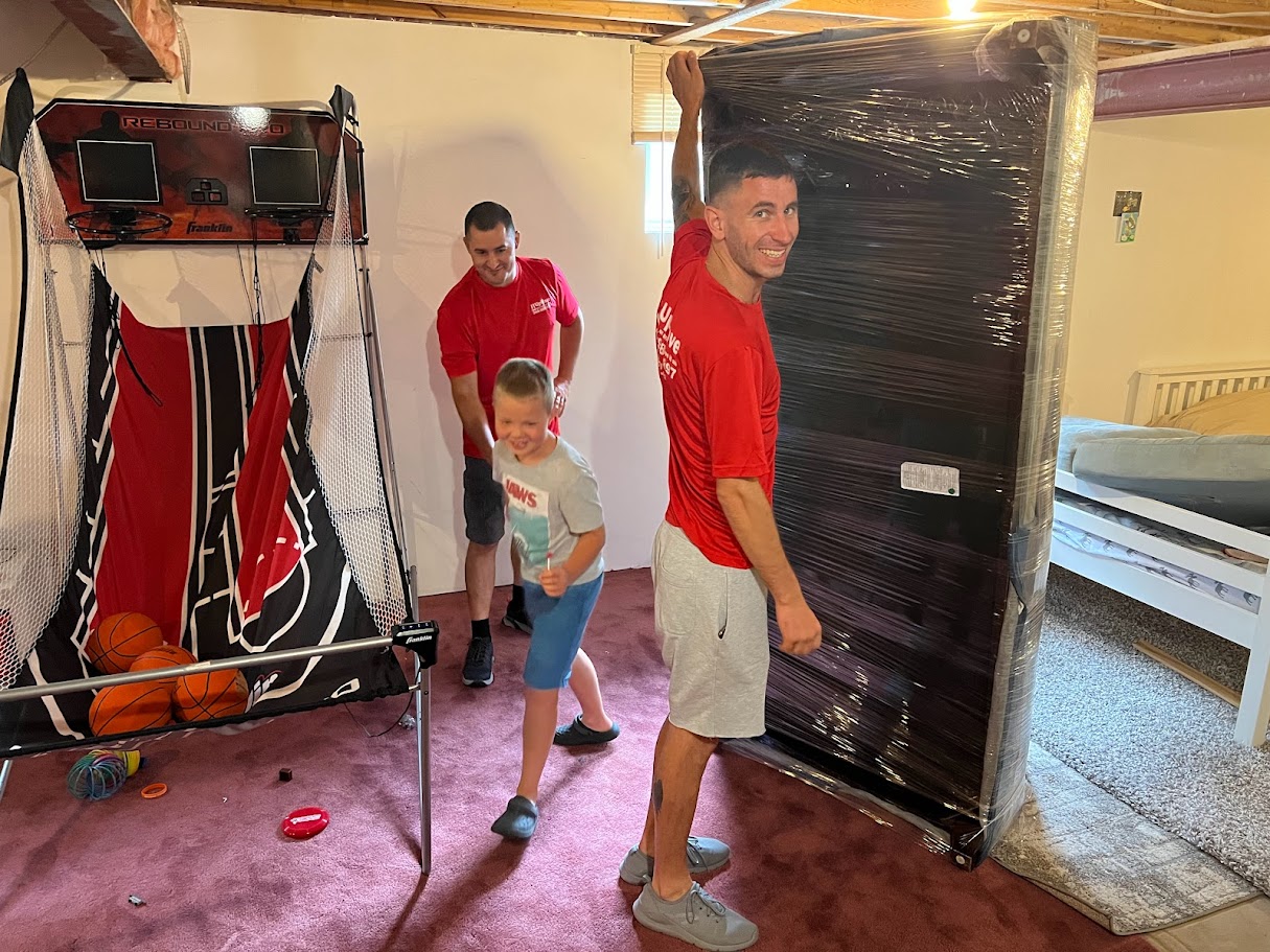 Two men in red shirts and a child in a grey t-shirt in a basement, one man holding a large wrapped item, basketball arcade game and basketballs to the left.