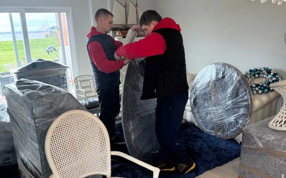 Two men wrapping and preparing furniture covered in plastic wrap inside a living room.