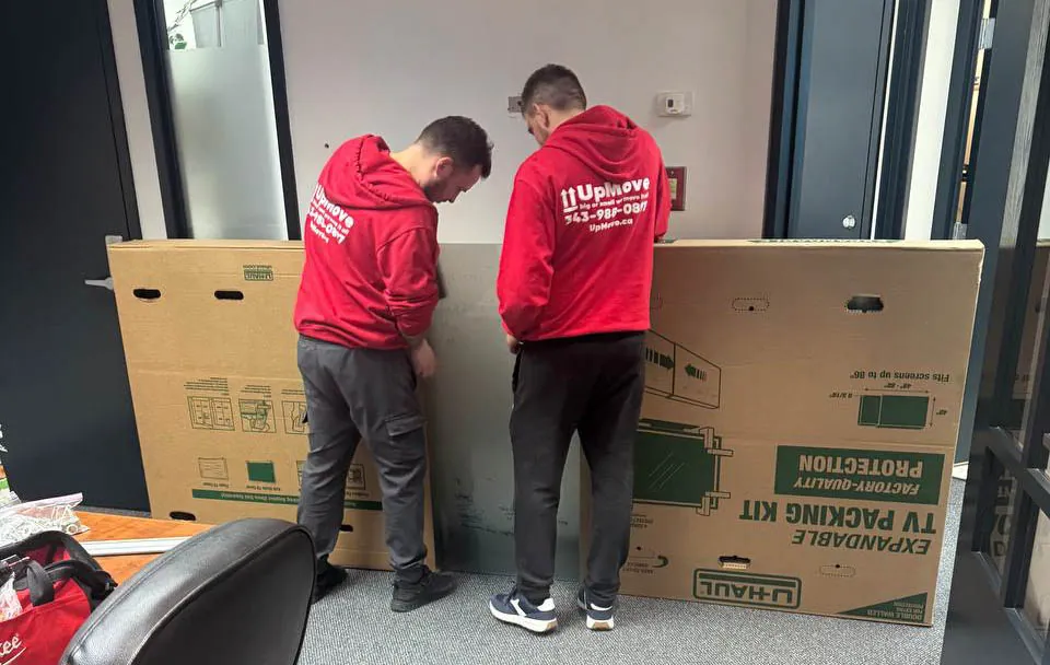 Two movers in red UpMove hoodies packing a flat-pack TV kit inside an office space.