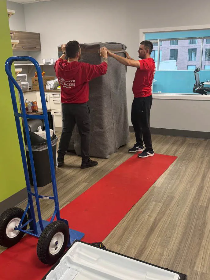 Two men in red shirts wrapping a large item with a gray moving blanket inside a room with wooden floors and a blue hand truck nearby.