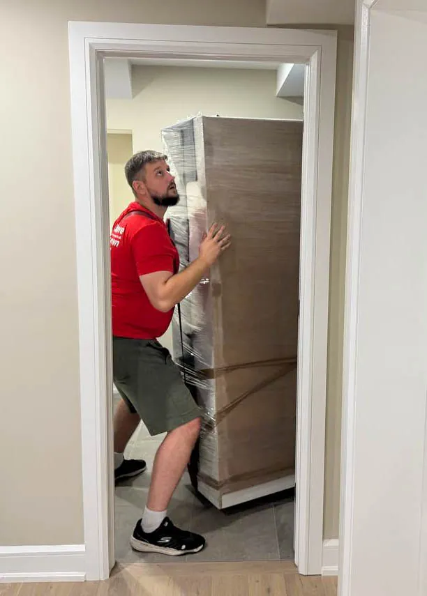 Man in red shirt and gray shorts carrying a large, shrink-wrapped piece of furniture through a doorway.
