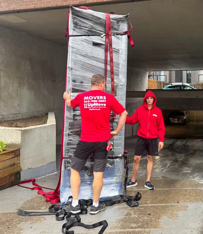 Two movers wearing red shirts secure a large wrapped piece of furniture with red straps near a parking garage entrance.