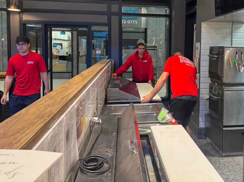 Three workers in red shirts arranging stainless steel and white countertops inside a commercial kitchen area.