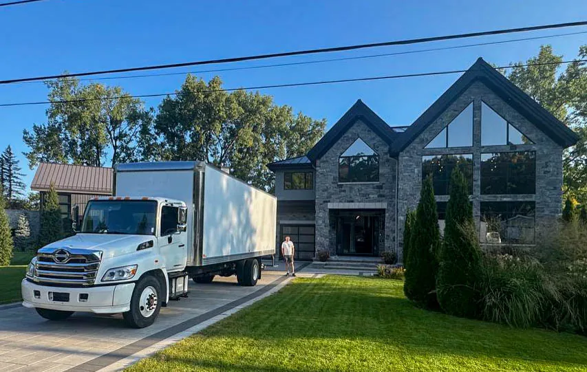 White delivery truck parked on driveway next to modern stone house with large windows and well-maintained lawn.