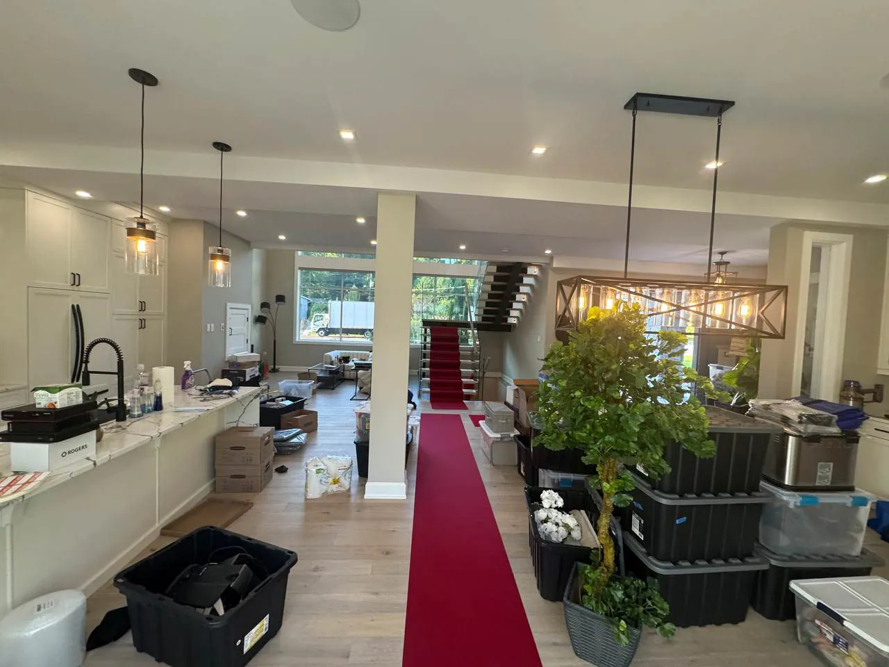 Spacious modern room with light wood flooring, a red carpet runner leading to a staircase, kitchen area with hanging lights, and various packed storage bins.