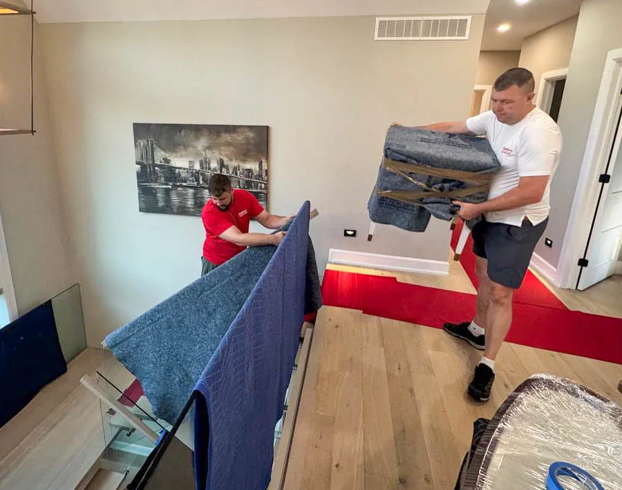 Two men moving furniture wrapped in protective blankets inside a modern home with wooden floors.