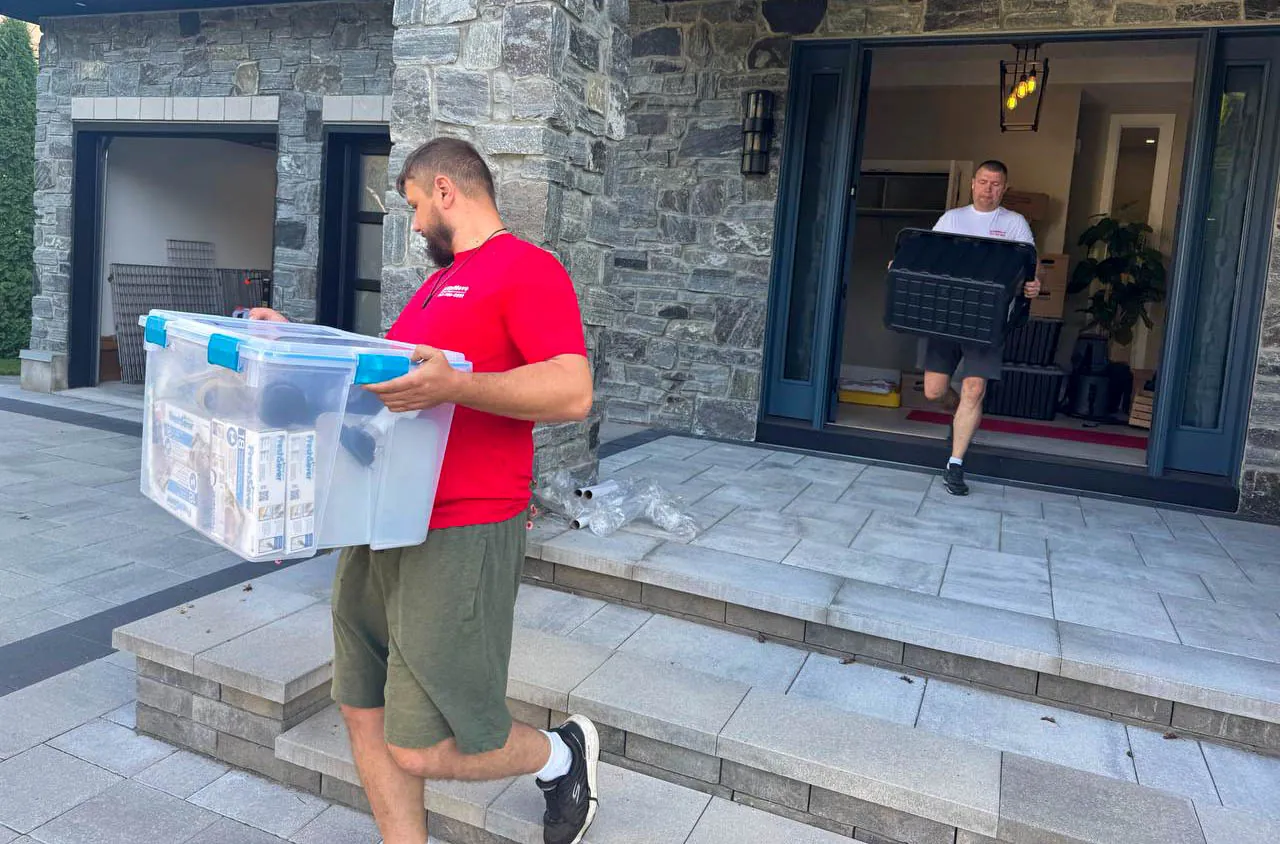 Two men carrying plastic storage containers outside a stone house with an open door.