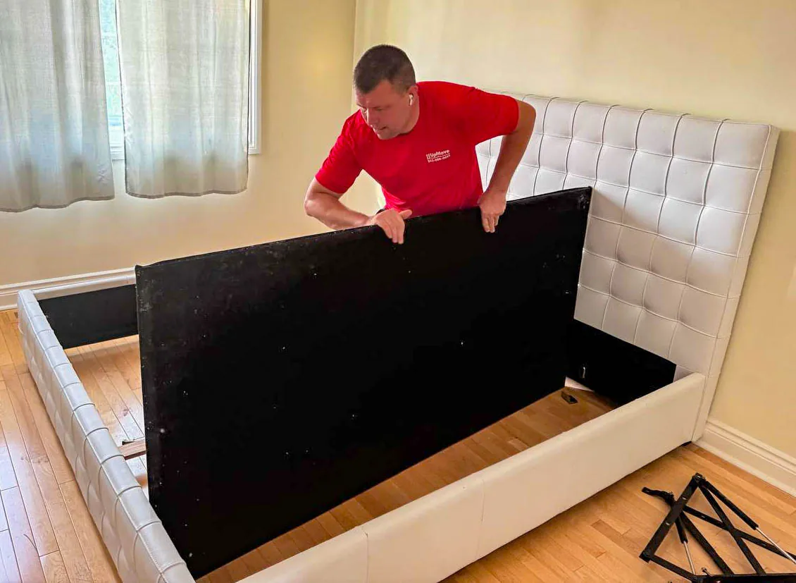 Man in a red shirt assembling a white upholstered bed frame in a room with hardwood floors.