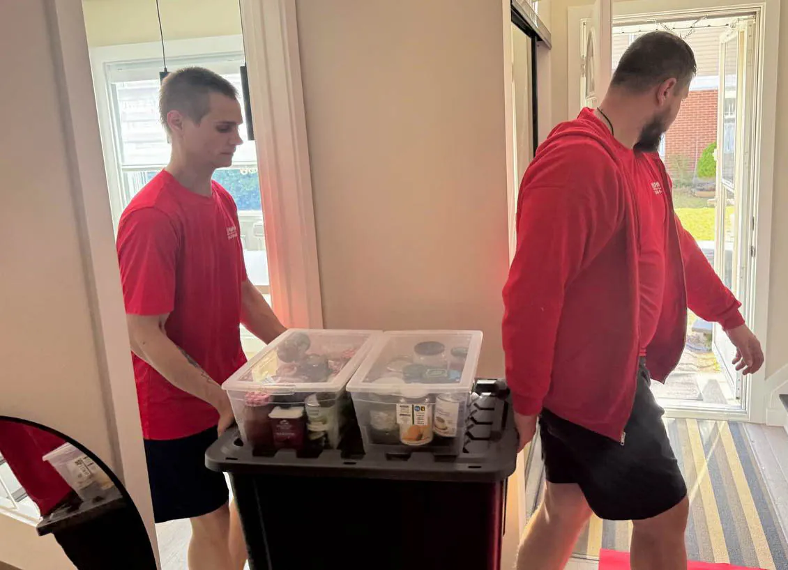 Two men in red shirts carrying a large black bin with two clear plastic containers on top inside a home near an open door.