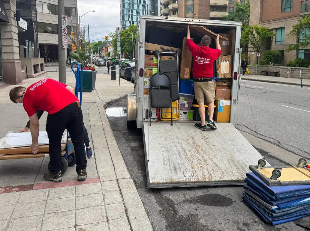Two movers in red shirts loading boxes and furniture into an open moving truck parked on a city street.
