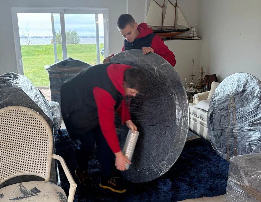 Two men wrapping large round furniture pieces with plastic wrap inside a room with a window showing grass and water outside.