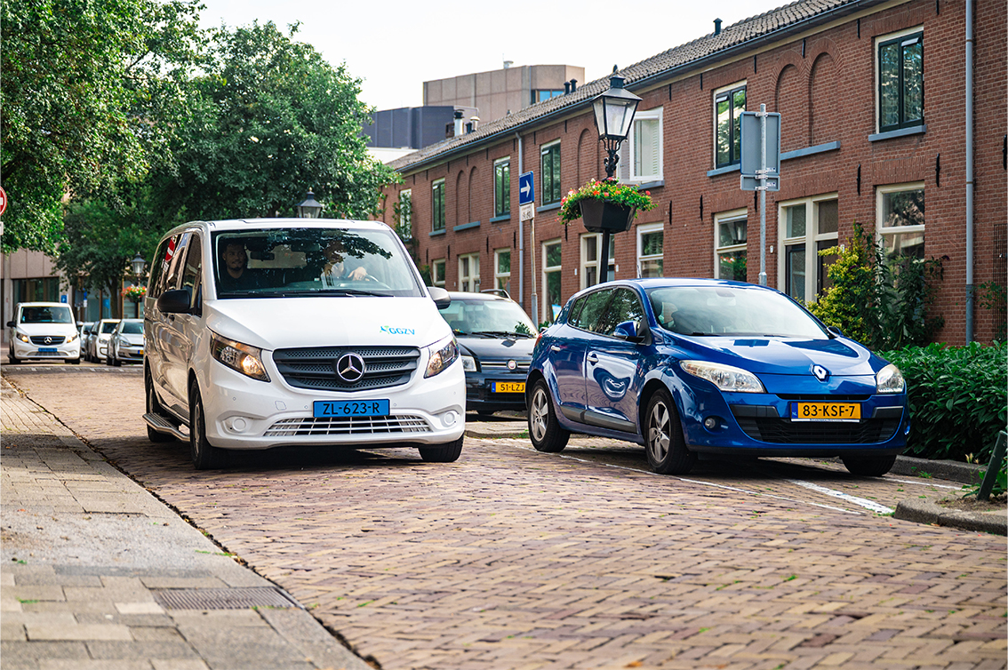 Witte bus en blauwe auto van GGZ Vervoersdienst in een woonstraat