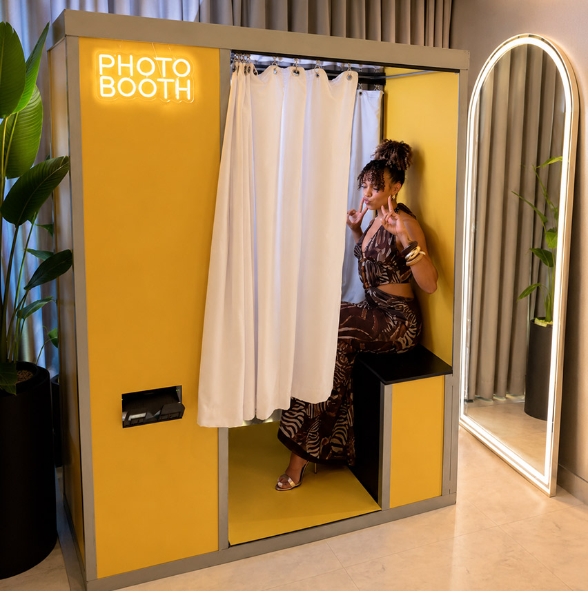 Woman in patterned dress posing inside a yellow photo booth with a white curtain and lit sign.
