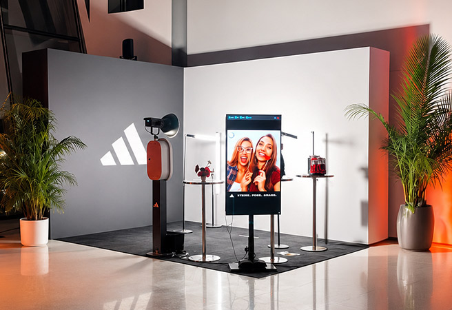 Modern photo booth with digital screen showing two smiling women, surrounded by plants and stands.