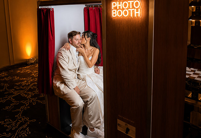Bride and groom in white attire sharing a tender moment inside a photo booth with red curtains.