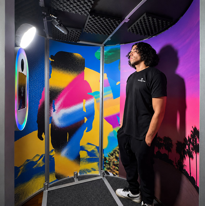 Man with curly hair in black outfit standing inside colorful photo booth with lighting and microphone.