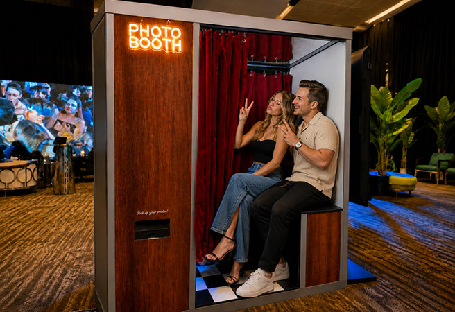 Two people posing and smiling inside a photo booth with a neon photo booth sign above.