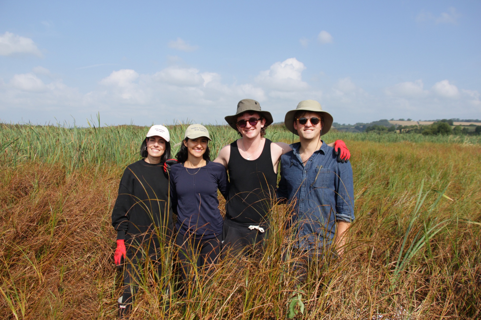 Four smiling people standing close together in a field of tall grass under a partly cloudy sky, wearing hats and sunglasses.