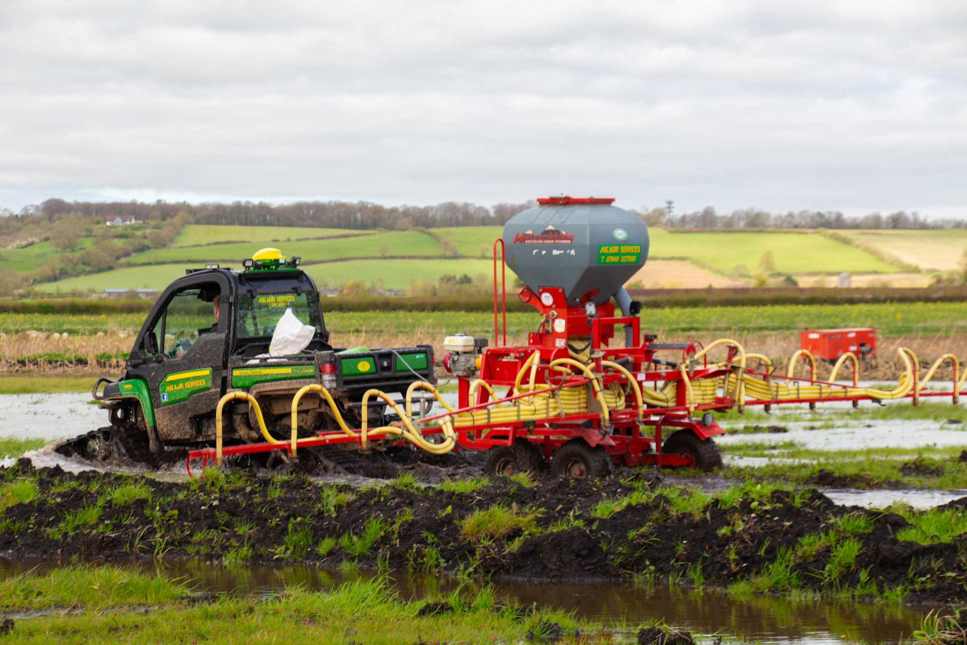 Green John Deere Gator vehicle towing red agricultural machinery in a muddy field with yellow tubes.