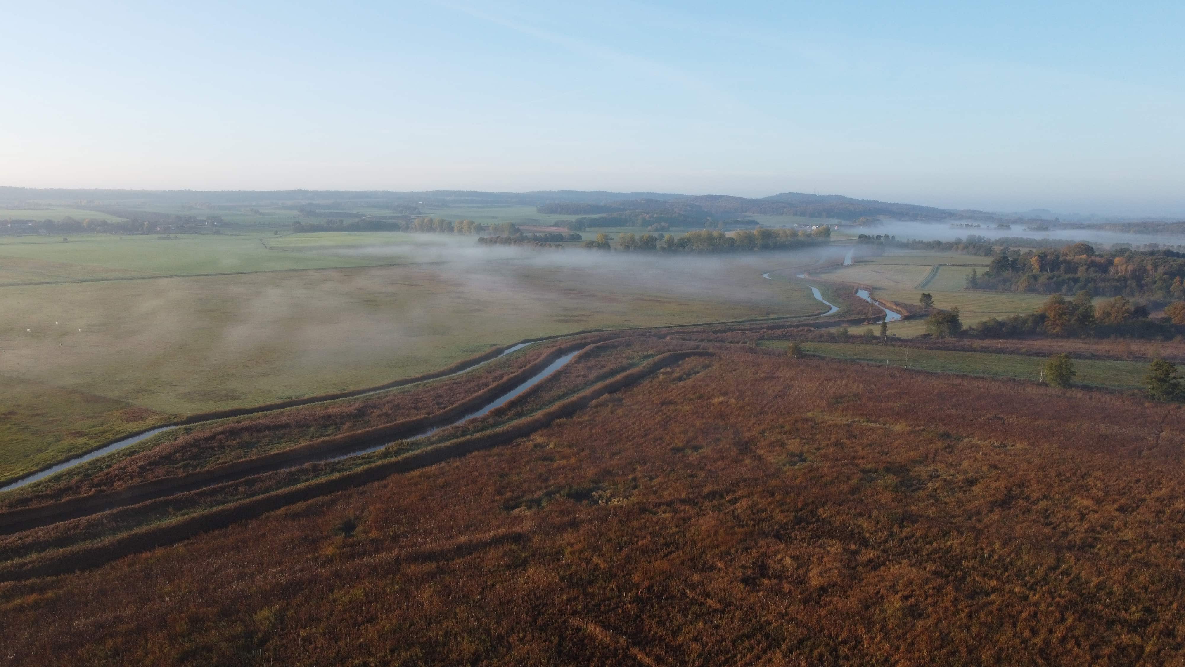 Aerial view of a misty rural landscape with winding waterways, green and brown fields, and trees under a clear sky.