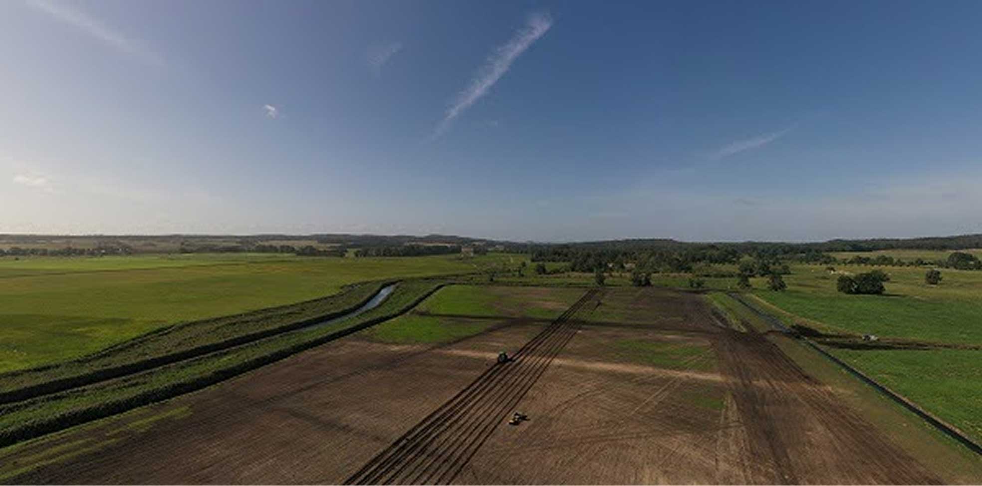Aerial view of farmland with a tractor creating parallel furrows under a bright blue sky.