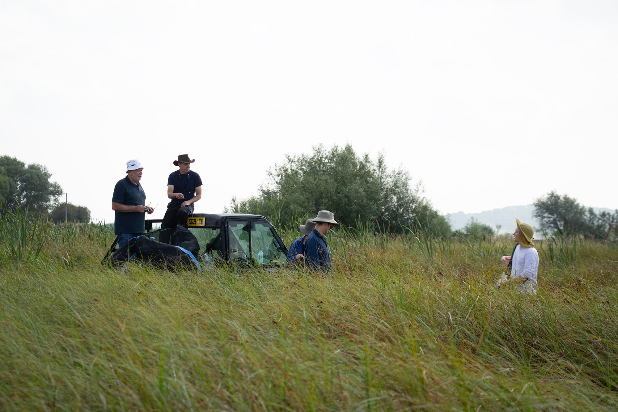 Four people wearing hats standing and sitting on an off-road vehicle in a grassy field with trees in the background.