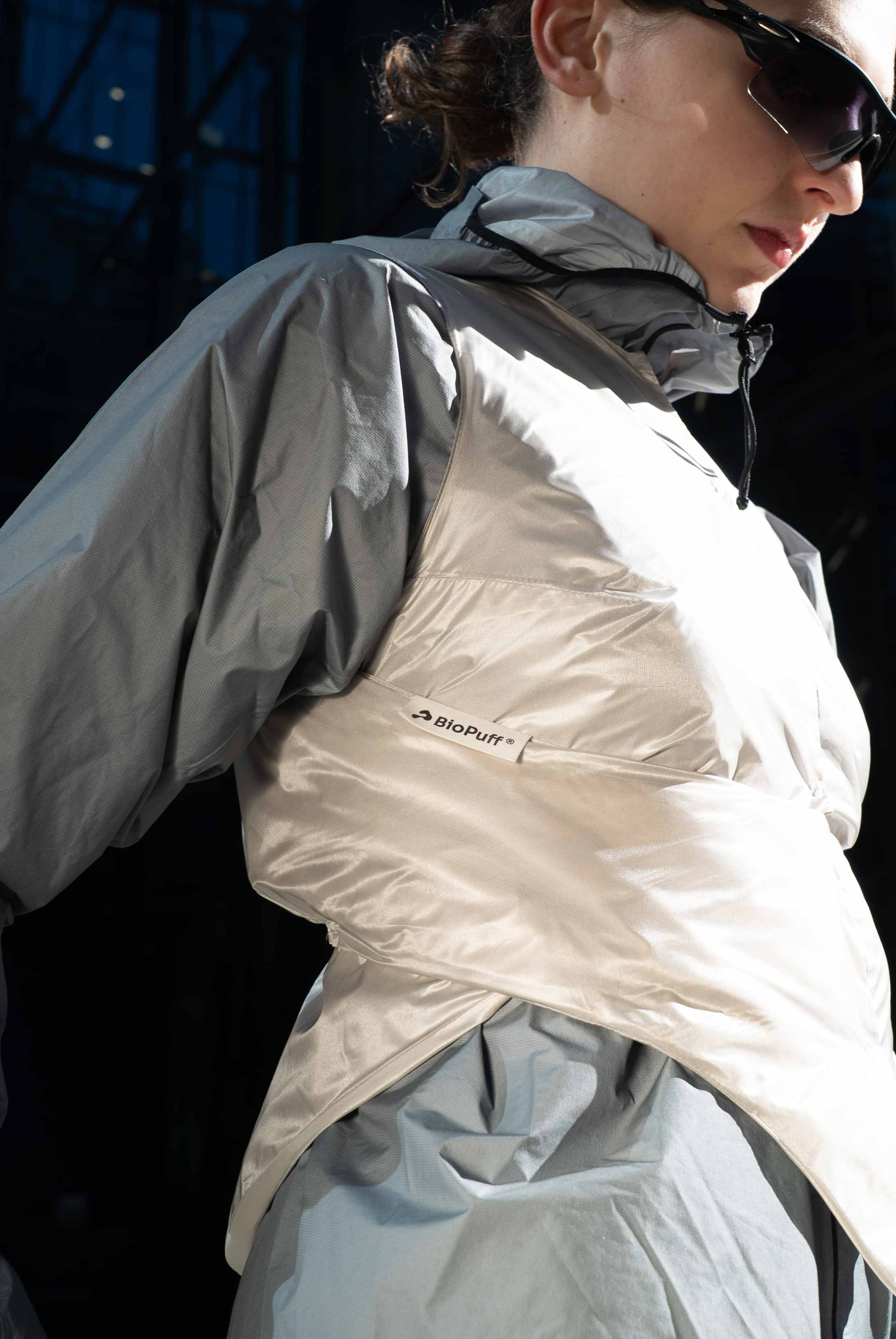 Close-up of a person wearing a reflective light beige BioPuff vest over a gray jacket and black sunglasses.