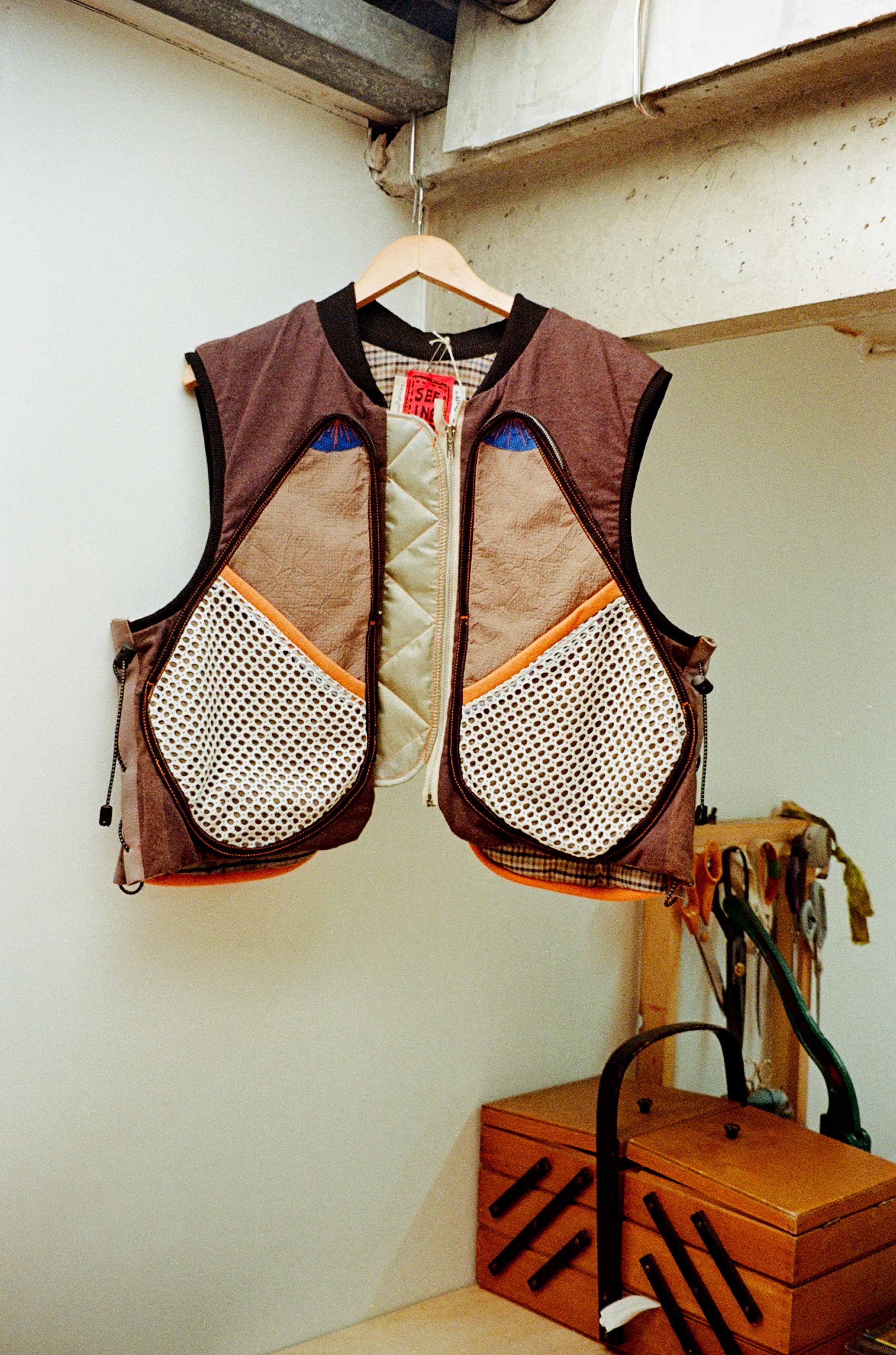 Colorful multi-fabric vest hanging on a wooden hanger in a workspace with tools and a wooden toolbox below.