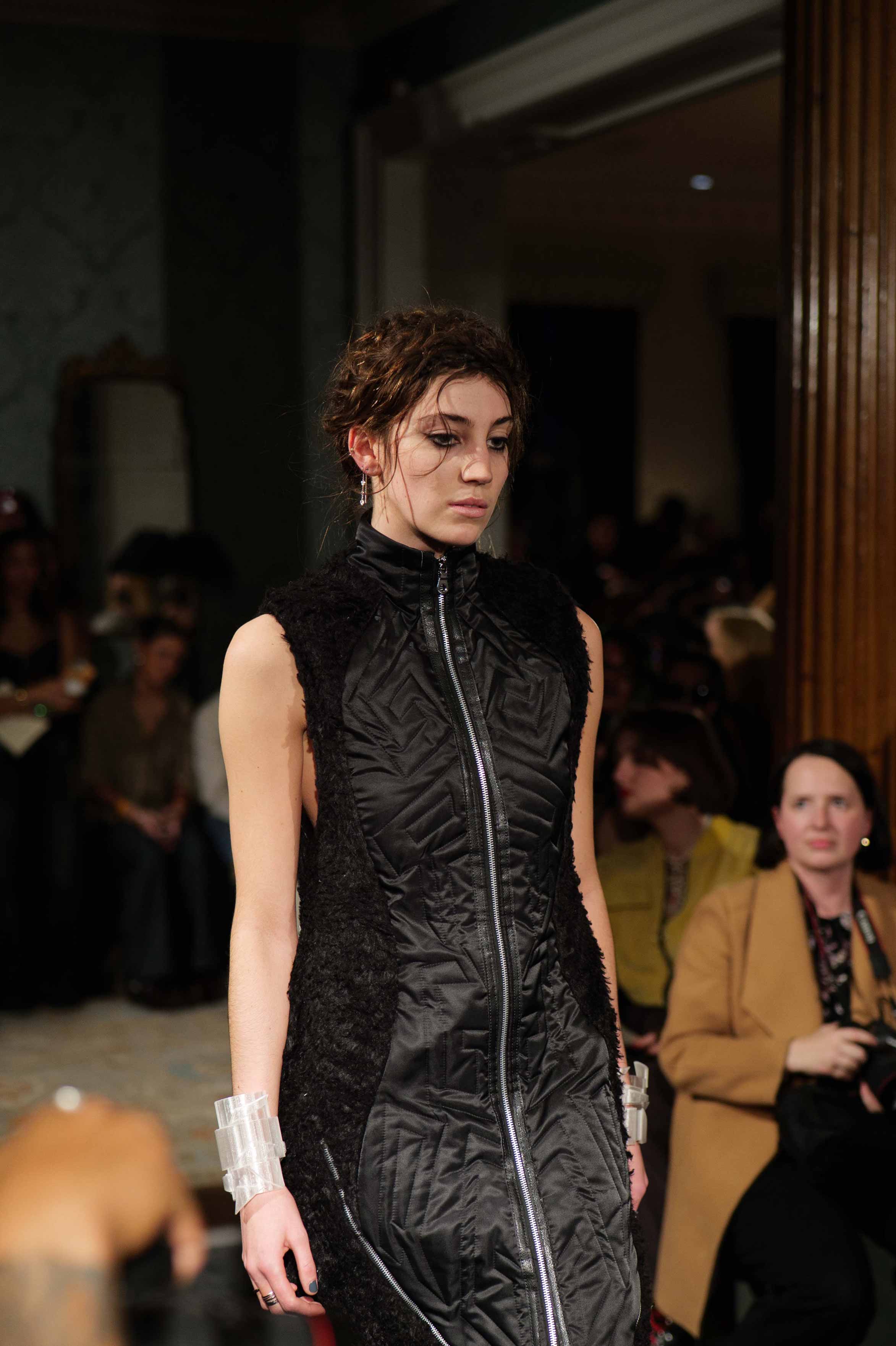 Female model walking runway in a sleeveless black dress with textured side panels and a front zipper.