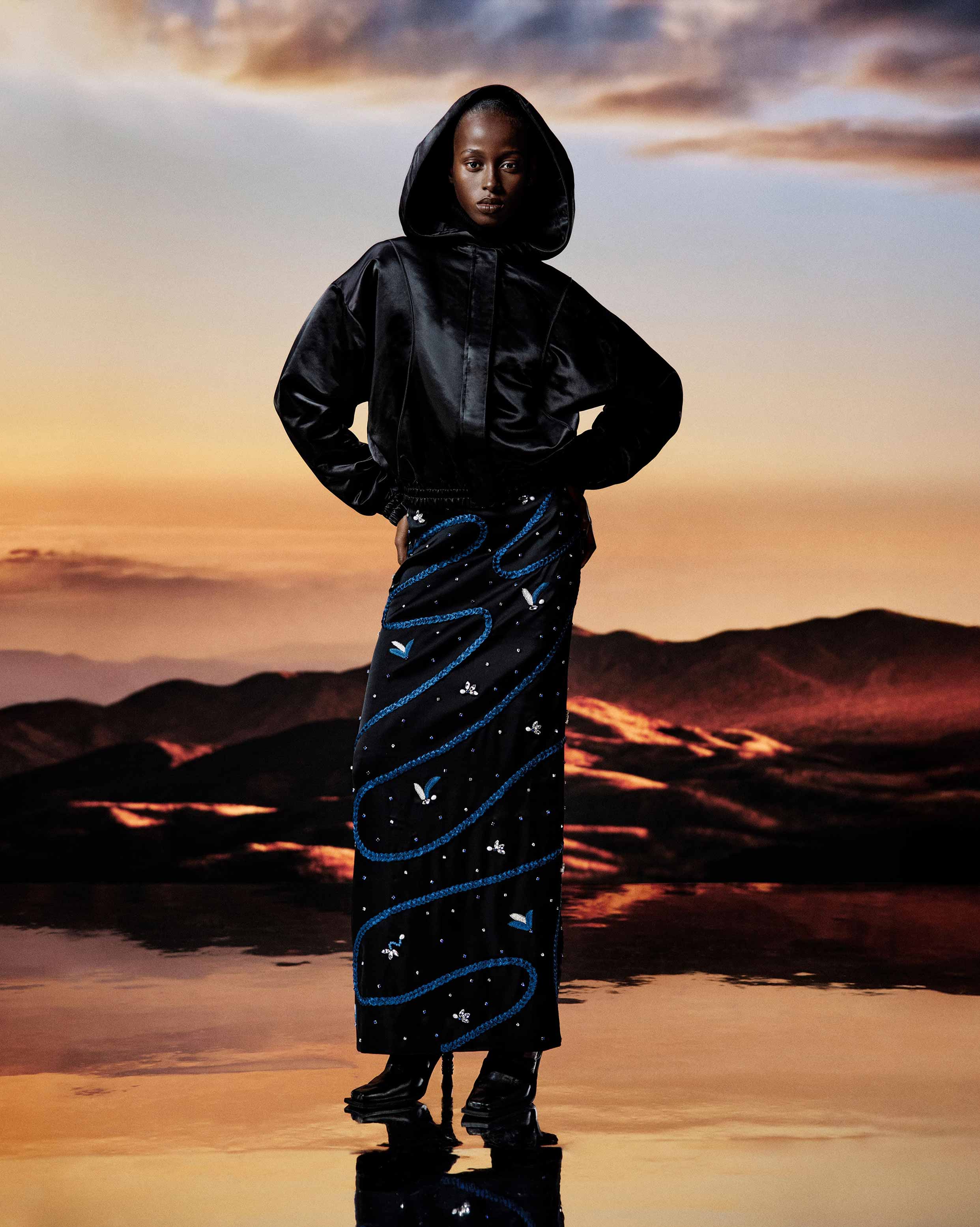 Model wearing a black satin hooded jacket and a long black skirt with blue swirls and small white floral patterns, standing on a reflective surface with mountains and a sunset sky in the background.
